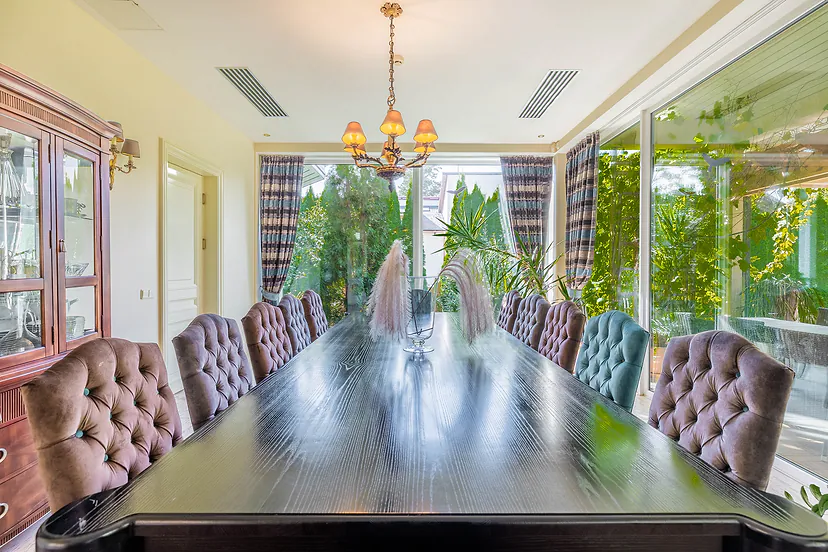 Dining room with a long table, upholstered chairs and a glass wall facing the garden