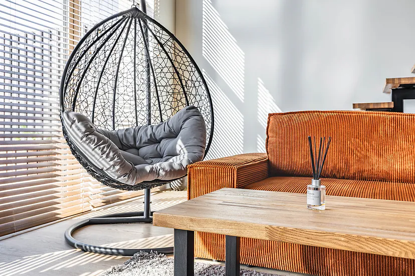 Pakabinama kiaušinio formos kėdė, oranžinė sofa ir medinis staliukas prie lango su žaliuzėmis