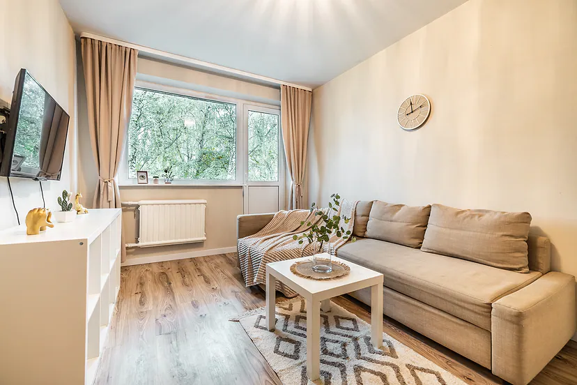 Šviesi svetainė su smėlio spalvos sofa ir užuolaidomis